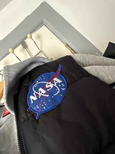 A black NASA jacket with a blue embroidered patch is hanging on a rack with other garments. The jacket is partially obscured by a grey hooded sweatshirt. The background appears to be a wall with a door and a hinge. The scene is indoors, and no specific location in Ballymena, United Kingdom is discernible.
