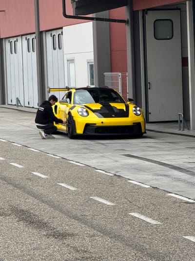 The image depicts a bright yellow Porsche 911 GT3 (992 generation) with black hood accents, a black roof, and a large black rear wing, stationary on a concrete surface in what appears to be a pit lane or garage area at the Autodromo di Cervesina, Cervesina, Italy. Its headlights are illuminated. A male individual, wearing a black hoodie displaying "ULL CAGNI Motorsport" on the back, dark trousers, and light-colored athletic shoes, is in a crouched position next to the vehicle's front left wheel, appearing to inspect or adjust the tire or wheel assembly. In the background, a building features multiple white vertical-folding garage doors to the left, and a single grey service door with a small window to the right, all set against reddish-brown and white wall sections. Dark utility pipes are visible above the right door. The foreground shows an asphalt track segment marked with broken white lines.