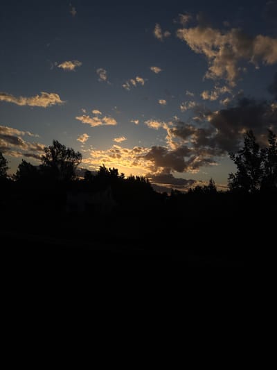 A vertically oriented photograph shows a sunset scene. To the left of the frame, a sky with blue hues is speckled with clouds that have their edges highlighted by the setting sun. The center features silhouetted trees and the suggestion of a structure or building, possibly a residence. To the right, the image is dominated by a field of black, indicating deep shadow or unlit space. The location is suspected to be somewhere in the Мажэйкаўскі сельскі Савет, Belarus. There are no observable people in this scene.