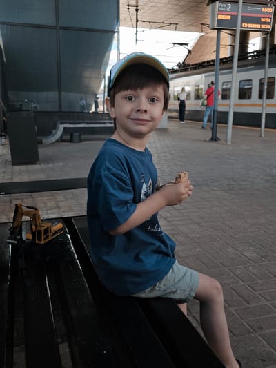 A young boy wearing a blue t-shirt and light-colored shorts sits on a dark wooden bench at a train station. He is wearing a cap with a colorful brim. In his hands, he holds a piece of food. To his left on the bench is a yellow toy construction vehicle. In the background, a train is visible at the platform, and a sign displays train information, including "Track 2". The station appears to be in Novoivanovskoe, Russia. The boy is looking towards the camera with a slight smile.