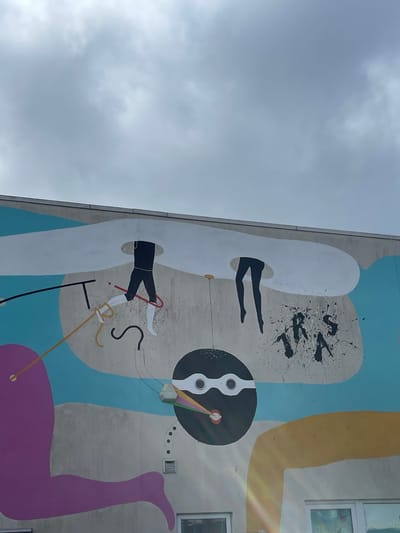 The image displays a vibrant, surreal mural painted on the side of a tall building, captured from a low, upward-looking angle against a cloudy sky. The artwork combines figurative elements with abstract shapes and colors.

On the left side of the mural, two separate pairs of stylized black legs emerge from elongated white, capsule-like forms. The upper pair of legs is pointed upwards, while the lower pair, clad in black pants and white socks/shoes, is more dynamic, holding a red cane or stick. Above the upper legs, black splattered paint forms what appears to be the letters "TRVS".

Towards the right and lower-middle sections of the mural, a prominent black circular shape with white internal details, resembling speakers or vents, is featured. A small, colorful, rainbow-like streak extends from its center. Large, flowing abstract shapes in blue, yellow, and purple frame the figurative elements, creating a dynamic backdrop. A notable large, purple, limb-like shape curves upwards from the bottom right of the mural. Various thin lines in black, yellow, and white are interspersed throughout the composition, adding to its abstract nature.

The building's concrete wall, which serves as the canvas, is a light grey. Two rectangular windows are visible on the far right edge of the image. The sky above is uniformly grey with diffuse clouds, suggesting an overcast daytime. A faint lens flare is visible on the right side of the image, indicating the sun might be partially visible or positioned off-frame. The overall scene captures a piece of modern public art within an urban environment, located in Portsmouth, United Kingdom.