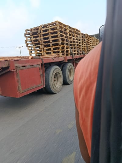 The image depicts a red flatbed trailer truck in motion on an asphalt road in Igbogidi, Nigeria. The trailer is heavily loaded with numerous stacks of light brown wooden pallets, secured by dark straps. Two axles, each with dual tires, are visible on the left side of the trailer. The road surface shows motion blur and faint yellow markings. A utility pole with wires is visible in the background on the left. The sky is light blue with some cloud cover. In the immediate foreground on the right, a partial view of a person's left shoulder and arm, clad in an orange shirt, is visible, alongside a dark vertical component of the vehicle from which the photograph was taken.