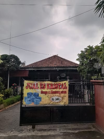 The image presents an outdoor scene in Kecamatan Gempol, Indonesia, under an overcast sky. The main subjects are a building, a metal gate, and a commercial banner.
A building, possibly a residence or small business, is partially visible with a distinctive reddish-brown tiled roof, suggesting traditional Indonesian architecture. In front of this building, a dark metal gate, consisting of vertical bars and solid lower panels, secures the property.
A prominent, brightly colored yellow banner is attached to the metal gate. This banner advertises "JUAL ES KRISTAL," meaning "SELL CRYSTAL ICE," written in large red letters across the top. Below this, smaller text reads "Harga : 1 Kantong cuma" (Price: 1 bag only), followed by "Rp. 5000," indicating a price of 5000 Indonesian Rupiah. The banner also features graphics of several blue, transparent blocks, likely depicting ice cubes or packaged ice, along with a starburst design around the price.
Green foliage, including tree leaves and bushes, is scattered around the scene, providing a natural backdrop and foreground elements. Against the uniformly gray, cloudy sky, several power lines are visible, crisscrossing the upper portion of the frame. The overcast conditions suggest a cloudy day, making it difficult to determine the exact time beyond daylight hours. There are no people visible, and the scene depicts a static, quiet street view, highlighting a local business selling crystal ice.
