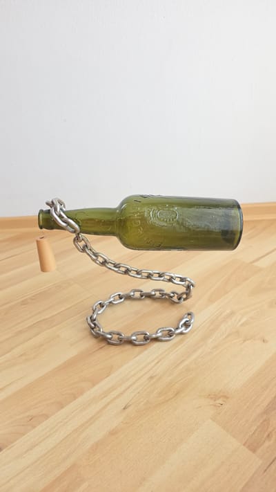 A green glass bottle is suspended horizontally in the air by a metal chain. A light-colored cork stopper is attached to the neck of the bottle by a string. The bottle is positioned over a light-colored wooden floor. Behind the bottle is a white wall. The image appears to be staged as an art piece or illusion. The image does not provide any information about the city of Crailsheim, Germany.