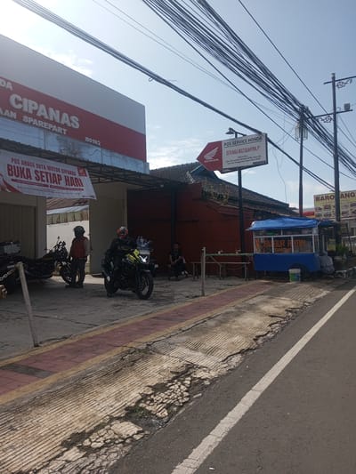 The image shows a street scene in Cipanas, Indonesia. On the left is a building with a sign that reads "CIPANAS AAN SPAREPART" and a banner that says "POS AHASS DUTA MOTOR CIPANAS BUKA SETIAP HARI". In front of the building, there are two motorcycles and two men standing. One man is sitting on a bench nearby. To the right, there is a "POS SERVICE HONDA" sign indicating "DUTA NIAGA MULTISEJAHTERA, PT". Next to the sign is a food cart labeled "BUBUR AYAM CIANJUR". A third sign is seen in the background for a "BARO ANG SEAFOOD & ANTARA" restaurant. Utility poles with numerous wires are visible against a partly cloudy sky. The road surface is cracked and worn, and a white line marks the edge of the road. The location is NOT Khe Luang, Laos.