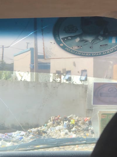 The image displays an outdoor urban environment in Basawa, Nigeria, viewed from within a vehicle through a cracked windshield. In the foreground, the vehicle's dashboard and windshield wipers are partially visible at the bottom. Two stickers are affixed to the upper right interior of the windshield; the prominent circular sticker shows a reversed portrait of a person in military uniform, with the mirrored text "GENERAL SANI ABAYCHA CCON" visible.

The midground features a large, irregularly shaped pile of mixed refuse, consisting of various plastic items, papers, and general waste, accumulated on the ground. Behind this refuse, a light-colored concrete perimeter wall, topped with strands of barbed wire, extends horizontally across the frame.

In the background, beyond the wall, multiple buildings with light-colored facades and blue trim are visible under a clear, bright sky. One building clearly shows several small, rectangular window openings. To the far left background, sparse vegetation and utility poles supporting power lines are present. No individuals are directly visible in the scene.