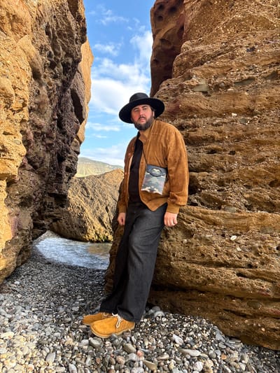 The image shows a man standing beneath a rocky overhang on a beach in Novyi Svet, Ukraine. He is wearing a black hat, a brown jacket, dark pants, and tan shoes. He has a small camouflage bag strapped across his chest. The beach is covered in pebbles and is next to water. The sky is visible through a gap in the rocks.