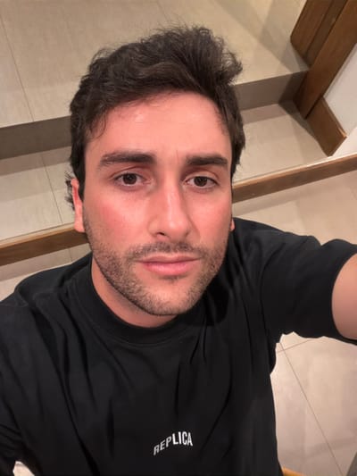 A man with dark hair and a short beard looks directly at the camera. He is wearing a black long-sleeved shirt with the word "REPLICA" printed on the right side. The background shows a tiled floor and wooden beams or panels. The image appears to be taken from a slightly high angle, with the man's face and upper torso in focus.