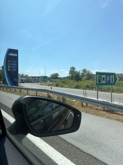 The image was taken from the interior of a vehicle traveling on a road in Općina Jakovlje, Croatia. In the foreground, a car's side mirror reflects the sky and surrounding landscape. To the left, a tall blue and white sign for the "INA" gas station displays fuel prices: 1.74, 2.09, 1.61, and 1.86. Beyond the sign, a gas station with multiple pumps and parked vehicles is visible. To the right, a green directional sign indicates services with icons for a gas pump, a cup of coffee, and an information symbol, pointing towards the right. The road is asphalt with white lines, and a metal guardrail runs alongside it. The environment is characterized by a clear blue sky with some faint contrails, green trees, and vegetation.