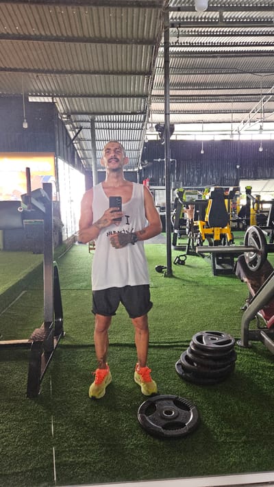 A man in a tank top and shorts stands in a gym, holding a phone and looking at the camera. The gym is located in Cam Ranh Ward, Vietnam. Weightlifting equipment and weights are visible. The floor is covered in artificial turf. The gym has a corrugated metal roof.