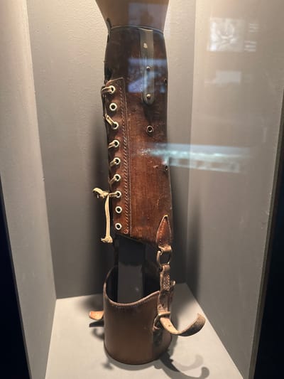 The image shows a close-up, side view of an antique leather prosthetic forearm. It is displayed in a museum setting, likely in a glass case as evidenced by reflections. The prosthetic is made of dark brown leather with visible wear and tear, suggesting age and historical use. It features a lacing system with metal eyelets and a fabric cord, similar to a boot, for adjustment and attachment. There are also metal studs or rivets along the length of the prosthetic, as well as a buckle and strap mechanism at the upper end. The prosthetic is mounted on a mannequin or a base, simulating it being worn. The background is a muted, neutral gray, which allows the prosthetic to be the focal point. Reflections on the glass reveal faint outlines of what appear to be exhibits or architectural features, hinting at the museum's interior. The lighting is somewhat dim, creating shadows that add to the artifact's historical feel. The overall impression is of a preserved piece of medical history, possibly from the late 19th or early 20th century, presented for educational purposes. Given the location context of Bern, Switzerland, this artifact could be part of a collection at a Swiss museum detailing medical history or the history of disability aids. There is no visible text in the image.