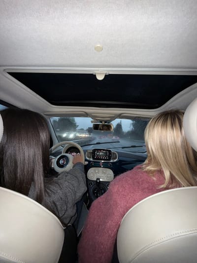 The image captures the interior of a car, looking forward from a rear perspective, at what appears to be dusk or night. The setting is on a road, likely urban or suburban, with other vehicles visible ahead, indicated by faint headlights and taillights. The windshield appears wet, suggesting rain or recent rainfall.
Two individuals are visible from behind. In the passenger seat, a person with long blonde hair, wearing a pink, fuzzy long-sleeved sweater, is partially seen. The driver, seated to the left, has long dark brown hair and is wearing a grey long-sleeved top. Their hands are on the steering wheel, which is white with a prominent "FIAT" logo in the center.
The car's interior features cream-colored upholstery on the ceiling and seats, contrasting with a darker dashboard. A rearview mirror reflects a faint image, possibly of an occupant. To the left of the central dashboard console, a mounted device, likely a phone or GPS, displays some text including "FM" and numbers such as "90.2", suggesting a radio is active. The car appears to have a sunroof, with a portion of its shade visible and partially open. The overall design details, including the steering wheel and dashboard elements, point to a Fiat 500 model.
The activity taking place is travel by car, possibly in traffic given the other vehicles on the road. The time of day is late evening or night, evidenced by the darkness outside, illuminated only by vehicle lights and streetlights. While not visually identifiable in the image, the provided context indicates the location is Villeneuve-Loubet, France.