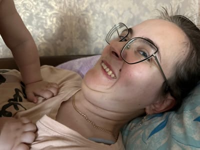 The image shows a close-up of a woman lying on her back, wearing glasses and a light-colored shirt. Her head is tilted back, and she is smiling. A baby's arm is visible in the upper left portion of the image, resting on the woman's chest. The background is a patterned wallpaper. The scene appears to be indoors, possibly a bedroom, given the presence of bedding and wallpaper. No specific location in Yazykova Pyatina, Russia, can be determined from the visual information provided.
