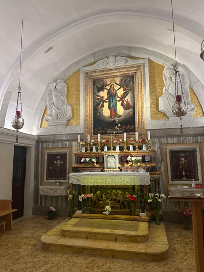This image presents a forensic view of a meticulously adorned religious chapel interior, located in Għarb, Malta. The central focus is an altar area against a wall dominated by a large, ornate golden-framed painting depicting the Virgin Mary in a blue and red robe ascending amidst numerous angels and cherubs, with figures looking upwards from below. This painting is set within an arched recess, backed by golden mosaic tiles and flanked by two large white marble sculptures of kneeling winged angels; the left angel has crossed arms, while the right's hands are clasped in prayer.

Below the painting, the multi-tiered altar features multiple gold-colored candlesticks holding white candles, interspersed with vases of red and white fresh flowers. A small silver crucifix and a small framed religious artifact are also on the upper tier. The mid-tier includes a carved or sculpted relief, likely depicting figures, flanked by vases holding white calla lilies and red flowers. The front of the altar is draped with a white lace cloth over a green fabric, displaying a repeating "MC" motif.

The vaulted ceiling is white, with a visible white rectangular panel, possibly a speaker or ventilation grill, on the left side. Two ornate silver-toned metal hanging lamps with red glass inserts are suspended by chains from the ceiling, flanking the central altar. The floor is covered in light brown tiles with an irregular, mottled pattern.

To the left of the altar, a dark wooden door with horizontal panels is partially visible, next to a simple wooden bench. Above this area, a framed reliquary with a red velvet background displays a crown-like object. Below it, a small wall-mounted shelf is covered with a white lace cloth, and a potted plant with purple and white flowers sits on the floor. To the right of the altar, a similar framed reliquary, also with a red velvet background, displays an intricate golden object. Adjacent to this, a wooden lectern holds an open book and a vase of pink flowers. A small pump bottle and a red container are also present on a nearby ledge.