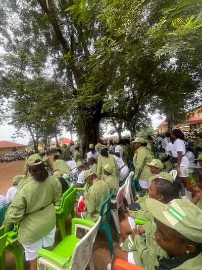 An outdoor event is underway in Ise-Ekiti, Nigeria, with a large gathering of young people, predominantly women, dressed in uniform. The uniforms consist of khaki shirts and shorts or skirts, and green caps with white and green stripes. Many are seated on brightly colored plastic chairs, while others stand or are partially visible in the background. The environment is characterized by a large, mature tree providing shade over the gathering, with other trees and buildings visible in the distance. The ground appears to be made of dirt. The overall atmosphere suggests a formal or semi-formal assembly, possibly related to a national youth service program or similar organization, given the matching uniforms. The lighting indicates it is daytime, with overcast or diffused sunlight filtering through the tree canopy.