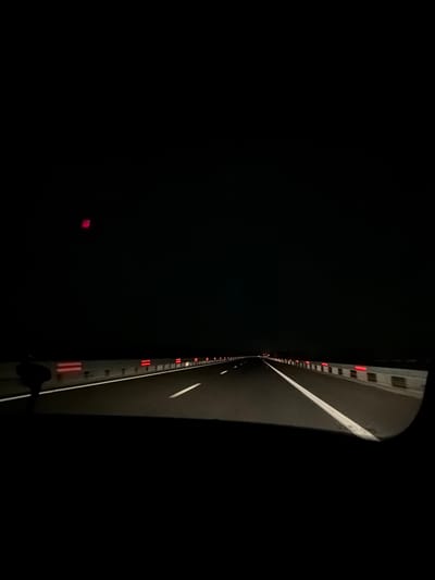 This image captures a nighttime scene on a road or within a tunnel in Chokoba, Bulgaria, viewed from what appears to be a moving vehicle. The composition is vertically oriented, with the illuminated path and a barrier predominantly on the right side, while a vast expanse of darkness fills the left and central areas.

On the right, a dark road surface is visible, marked by a distinct white line that functions as a lane divider or edge marking. Parallel to this road surface, a barrier or wall extends into the distance, adorned with numerous reflective red rectangular markers or small lights. These red elements provide visual guidance against the stark darkness.

The majority of the image, particularly the left two-thirds, is shrouded in deep black, emphasizing the nighttime setting and the absence of light in the surrounding environment. A faint, blurry red glow is discernible in the bottom-left dark area. The very edge of the frame on the far right shows a dark, curved element, consistent with the interior or side mirror of a vehicle, suggesting the photograph was taken from inside a car or similar transport.

The overall impression is one of travel through a dimly lit or dark environment, relying on road infrastructure for direction. There is no visible text in the image.