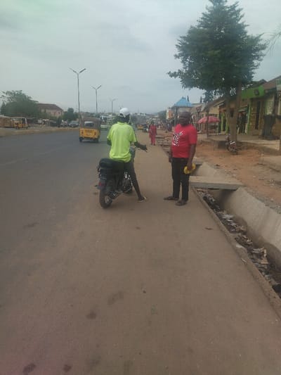 The image shows a street scene in Junction, Nigeria, at coordinates latitude: 8.958, longitude: 7.551. Two men are standing on the side of the road, one wearing a lime green shirt and a white helmet, and the other wearing a red shirt.  The man in the lime green shirt is standing next to a motorcycle and appears to be preparing to ride it. There is a concrete drainage ditch on the edge of the road. There are buildings on the opposite side of the road. The sky is cloudy and there are trees lining the road. In the background, three yellow vehicles, possibly tricycles, are parked and a few more cars are driving on the paved road.  The scene appears to be on a relatively quiet residential street in Junction.