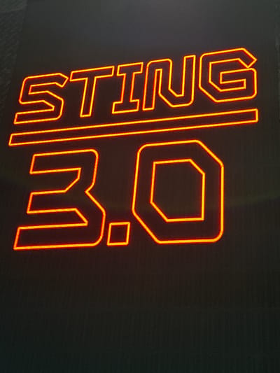 The image displays a neon sign with the text "STING 3.0" in orange and red hues against a dark, textured background. The sign is composed of blocky, stylized lettering. The word "STING" is positioned above a horizontal line, and below this line, the numerals "3.0" are displayed. The "3" is a large, stylized character, and the "0" is depicted as an octagon. The neon lights are bright, creating a glow that illuminates the surrounding darkness. There are no people or discernible environmental features that would suggest a specific city or location. The provided crops offer closer views of individual letters and parts of the numerals, highlighting the neon construction and design of the sign.