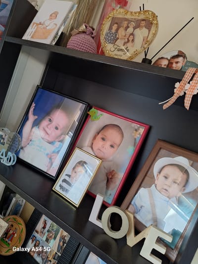 The image displays a detailed view of three shelves within a dark-colored shelving unit, likely located in a residence in Xylofagou, Cyprus. The shelves are filled with personal mementos and framed photographs, predominantly featuring infants and children.

On the top shelf, from left to right, a stack of paper items includes a photograph of an infant in a wicker basket. Adjacent is a small, purple, crocheted pouch with a gold ribbon. Next, a heart-shaped, clear resin photo frame with embedded gold flecks showcases a family portrait of five individuals (two adult males, one adult female, one child, one infant), inscribed with "Family" and the date "17.09.2023". To its right, a dark diffuser stick protrudes from a black base. Partially visible on the far right is an irregular-shaped decorative item featuring two faces and Greek text. Two checkered orange and white ribbon decorations with small white clover-like shapes hang above the shelf.

The middle shelf presents a collection of framed photographs. A clear, heart-shaped "DAD" ornament is partially visible on the far left. A black-framed photograph captures a smiling infant in a blue-accented top with a hand raised against a blue background. Next to it, a smaller gold-framed photo shows a young child in a striped shirt. A red-framed photo displays an infant lying on a floral background. On the right, a brown-framed photo features an infant in a white shirt, suspenders, and a white fedora-style hat. In the foreground of this shelf, four wooden letters spell "LOVE", with varying light and gold tones.

The bottom shelf is partially visible, showing a round, painted wooden plate depicting figures in traditional attire, with "CYPRUS" text visible. Next to it is a black-patterned photo album or large frame displaying multiple smaller photographs of adults and children in various settings. A "Galaxy A54 5G" watermark is present in the lower-left corner of the image.