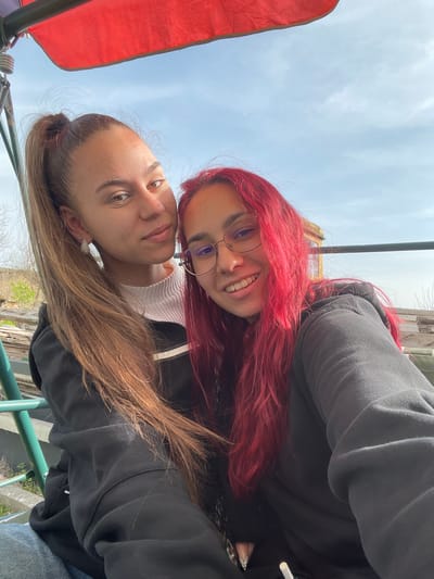 Two young women are close together, looking directly at the camera in a selfie. The woman at the top has bright red hair and wears glasses. The woman at the bottom has brown hair pulled back in a bun. Both are wearing black hoodies. The background shows a partial view of a blue sky and what appears to be a green metal railing and foliage, suggesting they may be on a bridge or elevated platform. The location is likely Muselievo, Bulgaria. A red fabric shade is present. The image suggests an outdoor setting during daylight hours.