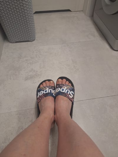 A pair of feet wearing black slides with "Superdry" text in white and a floral pattern. The floor is tiled and grey. A wicker laundry basket is on the left, and part of a washing machine is visible on the right. The scene is indoors.
