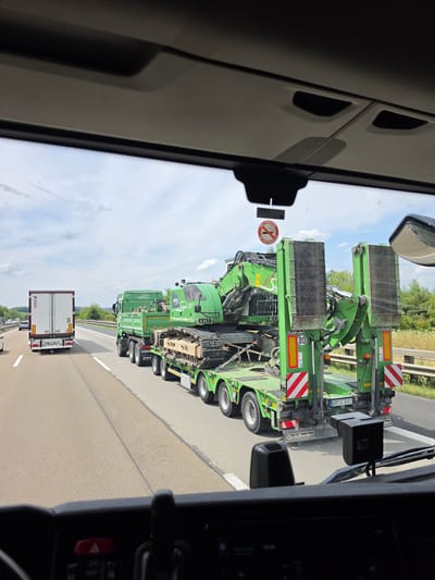 The image, taken from the perspective of a vehicle driver in Görgeshausen, Germany, shows a highway scene. The primary focus is a large green excavator being transported on a low-loader trailer. The trailer is attached to a green truck with a tipper body. The excavator is secured to the trailer with chains and straps. Both the truck and trailer are marked with safety reflectors and hazard warnings. The license plate on the trailer is visible as "BOR S 3081". In front of this convoy, a white refrigerated truck is traveling in the same direction. Other vehicles are visible in the distance on the highway. The environment is a sunny day with some clouds in the sky. The highway appears to be a multi-lane road with a grassy verge and trees lining the side. A "No Smoking" sign is affixed to the inside of the vehicle's windshield. The driver's dashboard is partially visible in the foreground.
