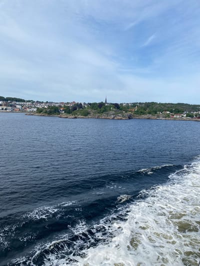 This is a vertical, medium-shot photograph taken from a boat moving through a body of water, likely a fjord or coastal area, near Larvik, Norway. The scene depicts a scenic view of a waterfront town nestled along a lush, green, and hilly coastline. The town is characterized by clusters of white and red-roofed buildings, with a prominent church steeple visible among them. The water in the foreground is dark blue, with white foam and waves indicating the boat's movement. The sky is a pale blue with wispy clouds. The time of day appears to be daytime, and the weather seems pleasant. The overall mood is serene and picturesque, highlighting the natural beauty of the Norwegian coast and its charming settlements. There is no visible text in the image.