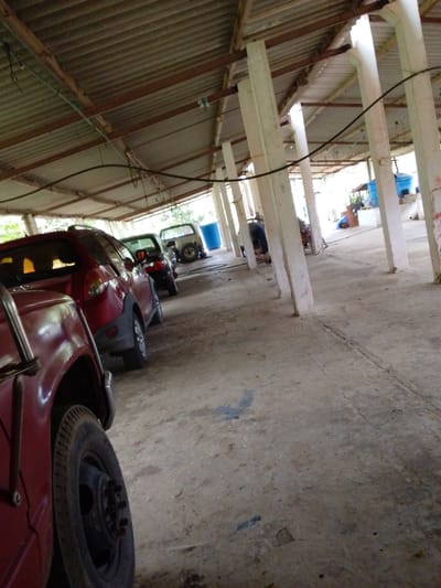 The image shows a row of cars parked under a covered structure. The structure appears to be an open-air shed with a corrugated metal roof supported by concrete pillars. The floor is concrete and appears worn and cracked in places. The cars are various colors, including red, black, and silver. One red car in the foreground has its front wheel visible. In the background, there are blue barrels and some building materials. The lighting suggests it is daytime, and the overall scene appears to be in a rural or industrial area. There is no visible text in the image. The setting is likely Atamo Sur, Venezuela, based on the provided location context. The image captures a still moment, with no activity or event clearly taking place beyond the parking of the vehicles.