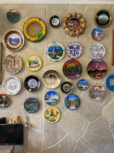 This media file showcases a collection of decorative plates and other trinkets mounted on a textured, stone-like wall. The items appear to be souvenirs, featuring images and text related to various travel destinations. Many of the plates are circular with colorful illustrations of cities, landscapes, and landmarks. Some examples include plates depicting "ARMENIA," "Lisboa," "ISTANBUL," "EGYPT," "PRAGUE," "BALI," "ANTALYA," and "amsterdam." There is also a circular item with shells surrounding a central image, labeled "Сухум."
Other objects include a sunflower-themed dish with bees and ladybugs, a brown-toned plate with a castle illustration, and a round plate with a mountainous landscape. A black circular item with a keychain attached displays a cityscape and the word "BAL." Further down, a blue circular item features buildings and palm trees, with text that appears to be "АБХАЗИЯ." A rectangular black screen is visible in the bottom right corner. The overall impression is of a personal collection of mementos, likely gathered from various travels. The wall's texture suggests an indoor setting, possibly a home or a display area. The lighting is consistent and appears to be artificial. There are no people visible in the image.