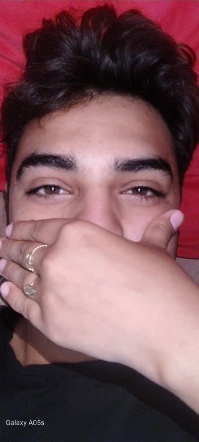 The image displays a close-up, rotated portrait of a young individual, likely male, lying down in an indoor setting. The subject's face is partially obscured by their right hand, which covers the lower portion, revealing only their dark brown eyes and prominent eyebrows. The eyes are looking directly at the viewer with a calm expression. Two rings are visible on the fingers of the right hand: a tear-drop shaped ring on the index finger and a thin, textured band on the middle finger. The individual has dark, wavy hair, visible against a red fabric background, suggesting a bed or couch. A dark, possibly black, garment is visible on the left side of the frame. The visible text "Galaxy A05s" is watermarked vertically on the top left of the image, indicating the camera used. The overall scene conveys a relaxed and candid moment. The location context is Zavoy, Bulgaria.