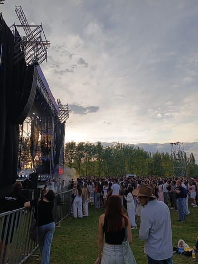 The image captures a lively outdoor concert or festival setting in Karajgach, Kyrgyzstan. A large crowd of people is gathered on a grassy field, facing a substantial stage with scaffolding and lighting rigs. The stage is draped with black fabric and adorned with numerous lights, some of which are illuminated and casting beams into the smoky atmosphere. On stage, performers can be seen, though they are not distinctly detailed. The time of day appears to be late afternoon or early evening, with the sun setting and casting a warm, soft light through the trees that line the perimeter of the field. The sky is partly cloudy, with a mix of grey and lighter patches.
The crowd is diverse, with individuals of various ages and attire, many holding up their phones to capture the event. A metal barrier separates the audience from the stage area, with some individuals leaning on it. One person in the foreground, to the left of the barrier, is wearing a black t-shirt with white text that reads "SOUND.KG Technical Staff". Another person, slightly further back and to the right of the barrier, is wearing a black t-shirt and jeans and is taking a photo with their phone. In the mid-ground, a man in a light blue striped shirt and a brown cowboy hat is looking towards the stage. The overall mood of the scene appears to be one of excitement and enjoyment, typical of a public gathering centered around music. The presence of trees and the open field suggest a park or a large outdoor venue.