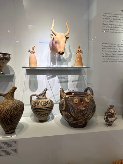 The image shows a display of ancient Minoan artifacts in a museum. The objects are housed in a glass-fronted display case with white walls and shelves. On a top shelf, there is a wooden sculpture of a bull's head and two small clay figurines of women. Below this, on a lower shelf, are several large pottery vessels. The closest vessel on the left is a tall, dark-colored vase with a pattern of intersecting lines. To its right is a lighter-colored jug with black painted decorations. In the center of the lower shelf is a large, bulbous pot with elaborate black and red painted designs, including circular motifs and figures. To the right of this, a smaller, dark-colored jug with white painted decorations is visible. A text panel is partially visible on the right side of the display case, and another text panel with Greek and English descriptions is placed on the lower shelf in front of the pottery. The overall setting suggests an archaeological exhibition. The text panel indicates that the artifacts are from the city of Heraklion, Greece, and are associated with organized cities, palaces, and maritime trade routes.