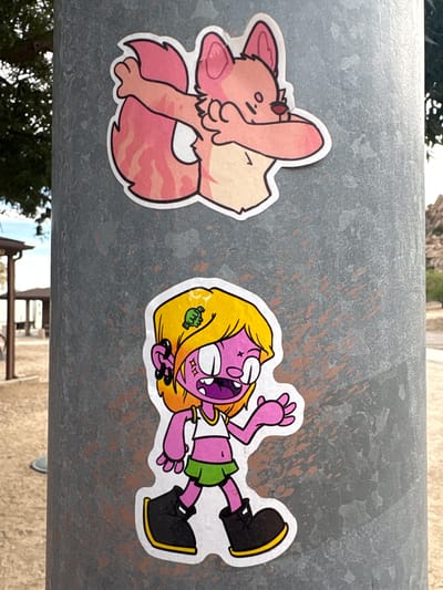 Two stickers are affixed to a gray metal pole in Dragoon, Arizona. The top sticker features a pink fox-like character with large ears and a fluffy tail. The character is in a pose with one arm bent and the other raised, covering its face. The bottom sticker depicts a cartoonish character with bright yellow hair, purple skin, and a wide, open mouth. This character wears a white crop top, a green skirt, and black boots with yellow trim. The background appears to be an outdoor setting with trees and a building visible in the distance.