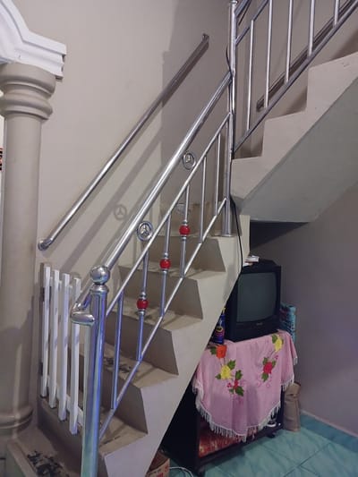 The image depicts a residential interior in Geudonggeudong, Indonesia, featuring a staircase with a stainless steel railing. The staircase is narrow and appears to be made of concrete. The railing is modern and includes decorative red spherical accents. To the left of the staircase, there is a white picket fence-like barrier and a decorative column. Below the staircase, on the right side, an older television set rests on a black cabinet covered by a pink checkered cloth with floral embroidery. Various objects, including a blue can and a rolled-up blue and white patterned fabric, are visible near the television. The walls are painted a neutral beige. The floor tiles are a light blue-green. The overall scene suggests a lived-in domestic space.