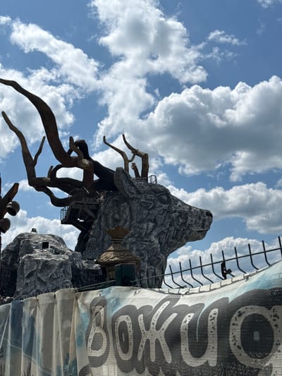 The image shows a monumental sculpture of a deer's head, intricately crafted from what appears to be stone and wood-like materials, against a bright blue sky with fluffy white clouds. The deer sculpture is part of a larger, rocky, mountain-like structure that also features what looks like a wooden tree or branch-like design extending from the top of its head. There are visible stairs and platforms built into the rocky structure near the deer's head, suggesting it might be an attraction or part of an amusement park. A black metal fence is visible in the foreground, and behind it, a draped banner or tarp with large, stylized, black lettering is partially visible. The lettering appears to be in a Cyrillic script, with parts of the word "ВОЖОД" (VOZHOD) discernible. The overall setting suggests an outdoor, potentially recreational, location. The image was taken in Ozernoe, Russia.