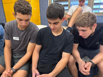 Four young males are depicted indoors in Ness Ziona, Israel. In the foreground, three males are seated, looking downwards. The male on the left wears a grey t-shirt with a white name tag displaying "נועם" (Noam) in Hebrew and a dark smartwatch on his left wrist. The central male wears a black t-shirt with a subtle circular logo and directs his gaze downwards, his hands positioned as if holding a small electronic device. The male on the right also wears a black t-shirt and a dark smartwatch on his left wrist, with his hands clasped and gaze fixed downwards. All three foreground individuals are wearing shorts. In the background, a fourth male sits on a light-colored ledge, possibly a window sill, with his right leg crossed over his left. He wears a white t-shirt, dark shorts, and dark socks, also looking downwards. The background on the left features a yellow wall with faint markings. To the right, behind the background individual, a window with horizontal blinds is partially visible. The scene suggests a shared moment of individual or collective focus, likely involving handheld devices, within a casual environment.