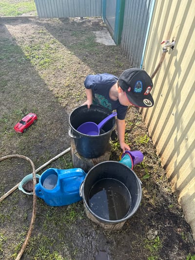A young boy, wearing a dark blue t-shirt and a black baseball cap adorned with Star Wars patches, is outdoors near a ribbed metal wall. He is in the process of playing with water. In front of him are several buckets and a blue watering can, some containing water. A red toy car is visible on the ground to his left. The ground appears to be a mix of dirt and grass, with some wet patches. A hose is also present, connected to a faucet on the wall. The scene suggests a casual outdoor activity, likely in a backyard or yard area. The location is specified as Chuy-Atasevo, Russia.
