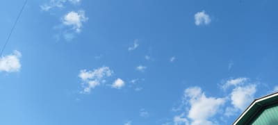 The image depicts a clear blue sky with scattered white cumulus clouds. In the bottom right corner, the peak of a green building with a white trim is visible, suggesting an outdoor scene. A thin, dark line, possibly a wire, extends diagonally from the top left edge of the frame. The scene is devoid of any people or specific actions. Based on the architectural style of the building and the overall ambiance, the image appears to be taken in the city of Kirsanov, Russia.