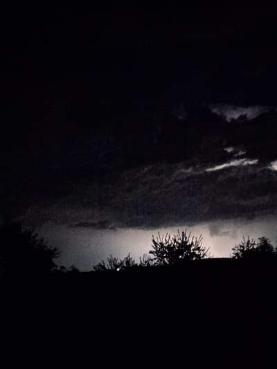 The image depicts a dark, silhouetted landscape at night, likely during a thunderstorm. A sliver of bright light, possibly lightning, illuminates the stormy clouds in the middle ground. The foreground is dominated by the dark outlines of trees against this backdrop. The scene appears to be an outdoor setting, given the presence of vegetation. The image's grainy texture suggests it was taken in low light conditions, possibly with a phone camera. There is no discernible location within Mlada Gvardia, Bulgaria, in the image itself.