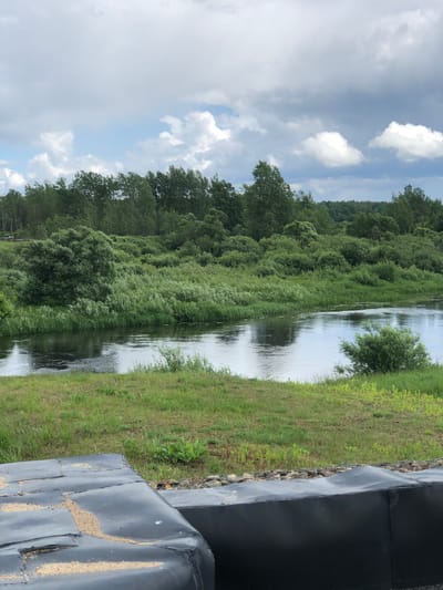 The image depicts a river flowing through a lush, green landscape. The foreground features a dark, textured surface that appears to be artificial, possibly part of a construction or embankment. Beyond this, a wide river meanders through the scene, reflecting the overcast sky. The riverbanks are densely covered with a variety of green vegetation, including trees, bushes, and tall grasses. In the background, a dense forest stretches across the horizon, with a slightly lighter shade of green suggesting a mix of deciduous and coniferous trees. The sky is filled with a dramatic display of clouds, with patches of blue sky visible between them. The overall impression is one of a natural, somewhat wild environment, possibly located in or near the city of Nadezhda, Russia, given the vegetation and cloudy sky typical of the region. There are no people or animals visible in the image.