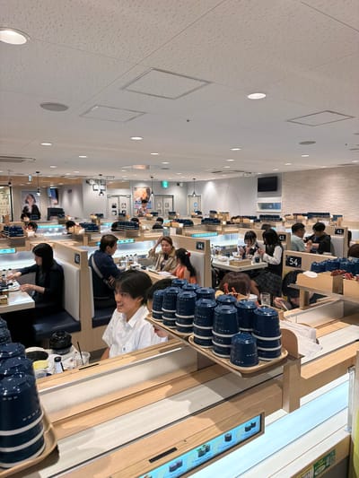 This is a photograph of the interior of a busy conveyor belt sushi restaurant located in Chiyoda, Japan. The perspective is from inside the restaurant, looking across a series of booths and a conveyor belt system.

In the foreground, on the right side, is a young man in a white collared shirt smiling and holding a tray stacked with blue ceramic cups. The tray is on a wooden shelf above the conveyor belt. To the left of the young man, also in the foreground, is a similar wooden shelf with more stacked blue cups. Below this shelf is a conveyor belt with several dishes of sushi visible, each with a label indicating its name and price. The conveyor belt runs through the center of the image, from the foreground towards the background.

The restaurant interior is characterized by light-colored wooden accents and blue booth seating. Several diners are visible in the booths. On the left side of the image, a woman with dark hair is seated at a table, facing away from the camera, and appears to be eating. Further back, in the center of the image, a group of people are seated in a booth, some of whom are interacting with the conveyor belt. More diners are visible in booths stretching into the background on both the left and right sides of the image. The ceiling is a typical drop ceiling with recessed lighting and ventilation panels. The overall atmosphere appears lively and crowded, consistent with a popular dining establishment. The signage on the wall includes Japanese text.