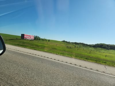 The image, taken from a vehicle traveling on a paved road in Hanley, Canada, captures a rural landscape under a clear blue sky. The foreground features the asphalt road with a white line marking the lane. To the right of the road, a grassy hill slopes upwards, dotted with several wooden fence posts. In the middle ground, a trailer with a red and white advertisement is parked on the hill. The advertisement displays text that reads "THE CANADIAN BREWERY" and "COMING SOON," along with stylized figures of people. The background consists of a line of trees and more rolling green hills under the expansive sky.
