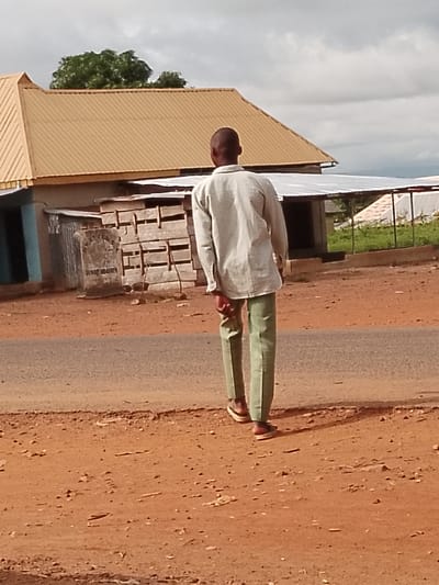A person is walking away from the camera on a dirt road in what appears to be the city of Ichol, Nigeria. The person is wearing a light-colored, long-sleeved shirt and light green pants. They are also wearing brown shoes. The background shows a building with a tan, corrugated metal roof, and other structures with similar roofing. There are trees and vegetation visible in the distance, and the sky is cloudy. The ground is covered in reddish-brown dirt with some scattered stones. A stone monument with some visible text, likely a historical marker, is to the left of the main building.