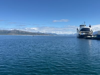 The image captures a serene waterscape at Lødingen, Norway, under a bright, partly cloudy sky. The scene is dominated by a large, modern ferry docked at a pier on the right side of the frame. The ferry is predominantly white and black, with its distinctive open bow facing the viewer, suggesting it's either docked or preparing for a journey. A small boat is visible in the distance on the left, cutting through the calm blue water and leaving a faint wake.
In the background, a range of majestic mountains forms a dramatic backdrop. Their slopes are covered in vegetation, and the peaks are partially snow-capped, hinting at the time of year or the high latitude. The clear blue sky is dotted with fluffy white clouds, indicating fair weather conditions. The water itself is a deep, tranquil blue, with gentle ripples reflecting the sunlight. The overall impression is one of peaceful natural beauty combined with the presence of human activity through the ferry and the dock infrastructure. The time of day appears to be late morning or early afternoon, given the brightness of the sun and the clear visibility. There is no visible text in the image.