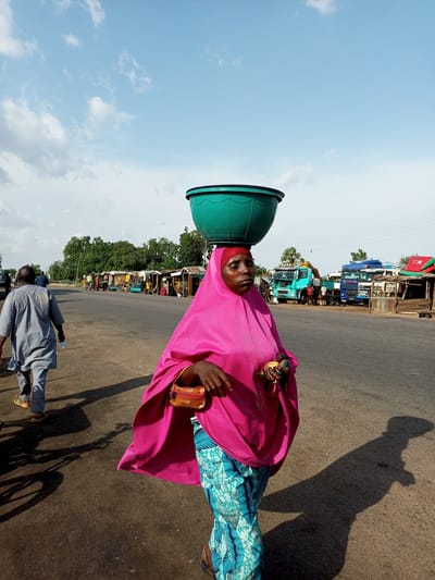 A woman with a large green bowl balanced on her head walks on the right side of a paved road in Gadao, Nigeria. She wears a pink hijab and a brightly patterned blue and turquoise skirt. A small, tan purse is clipped to her waist. Her right hand is raised, holding what appears to be a mobile phone. She casts a long shadow on the road. In the background, a man walks away from the camera on the left side of the road, carrying a water bottle. Across the road, a row of small market stalls and parked trucks are visible. The sky is blue with scattered white clouds.