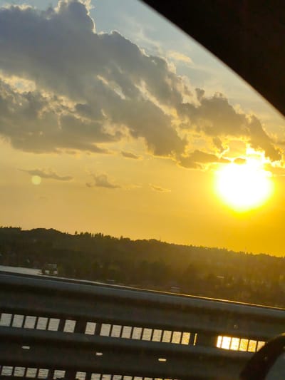 The image shows a sunset over a body of water, likely a lake or river, as viewed from a bridge. The sun is bright and partially obscured by clouds, creating a golden sky. Below the horizon, there's a tree-lined hill. A guardrail with rectangular openings lines the bottom of the frame, reflecting sunlight on the water visible through the openings. A portion of the car's interior is visible on the right and upper right of the frame. The location is identified as Пальники, Russia.