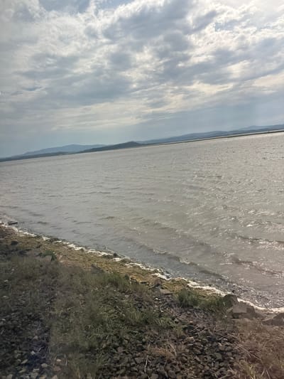 The image depicts a body of water with a rocky shore and a cloudy sky. The water is calm with gentle ripples, reflecting the overcast sky. The shore is composed of various sized rocks and some sparse vegetation. In the distance, a thin strip of land is visible, with faint outlines of hills or mountains. The sky is filled with a mix of dark and light clouds, suggesting a possibly overcast or partly cloudy day. The overall scene is natural and serene, with no discernible activity or human presence. The lighting suggests it could be daytime, though the cloudy conditions might obscure direct sunlight. The location is identified as Narbonne, France.