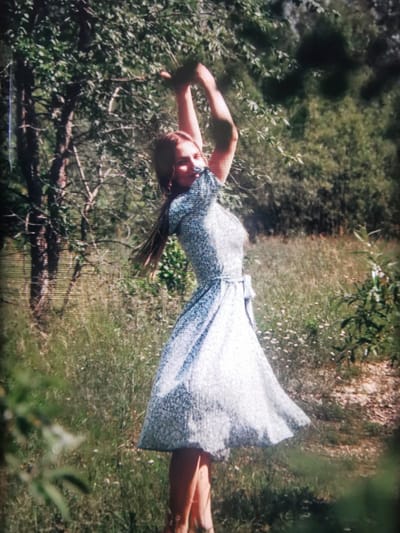 This is a photograph of a young woman in a light blue dress with a floral pattern. She is standing in a grassy field with trees and foliage in the background. The woman is captured in motion, with her arms raised above her head, appearing to be dancing or twirling. Her hair is long and dark, and she has a joyful expression on her face, with her head tilted back and eyes closed. The dress has a cinched waist with a tie and a flared skirt. The setting suggests an outdoor, natural environment, possibly a meadow or a park, on a sunny day given the lighting and shadows. The image has a slightly grainy quality, suggesting it may be an older photograph or taken with a less advanced camera. There is no visible text in the image. The scene evokes a sense of freedom, happiness, and connection with nature.