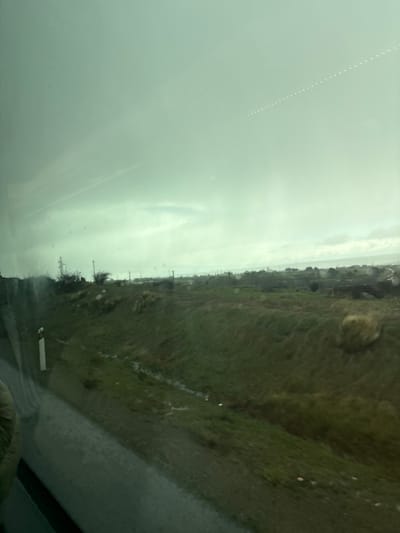 The image, taken from a moving vehicle, shows a view of a green, grassy hillside and a cloudy sky. The hillside is visible through the windshield, which is wet with rain. The sky is a muted green and is obscured by a layer of fog or cloud cover. Several small white objects are visible in the sky. The scene is likely taken near the coordinates 40.816, -4.192, which places the location near the town of La Losa, Spain.