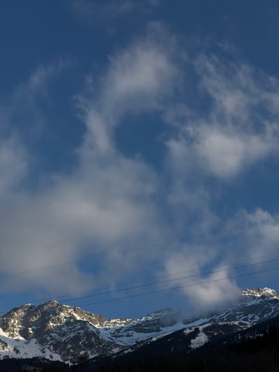 The image captures a daytime scene with a prominent mountain range partially covered in snow on the right side. The sky, filling most of the frame, is a clear blue with scattered white clouds. Several thin, dark, vertical lines, resembling power lines or cables, are visible in the center, running parallel to the mountain. The base of the mountain is shrouded in shadow or dark vegetation. Given the location hint of Saint-André, France, the mountain range is likely part of the French Alps.